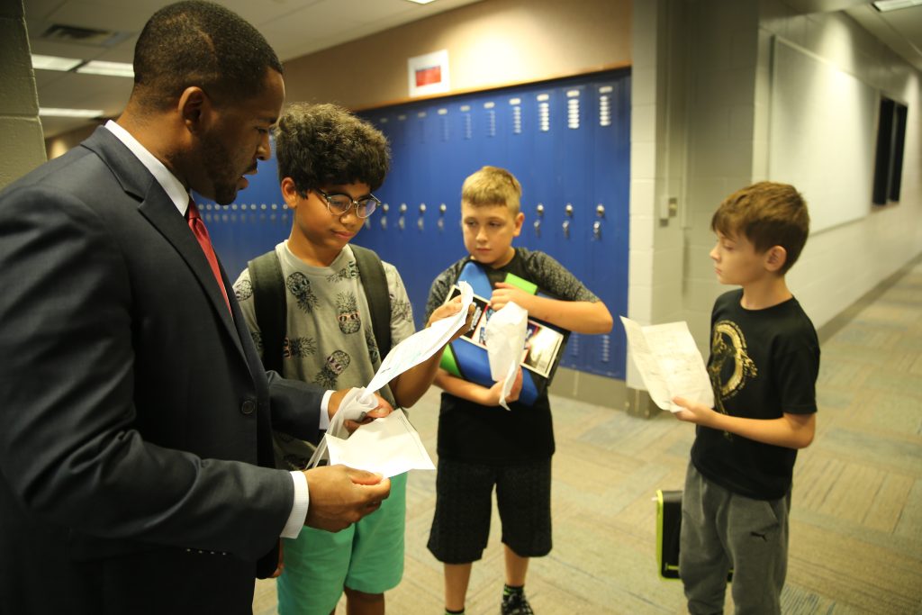 A man in a suit stands between two boys, handing them papers, while another boy looks on.